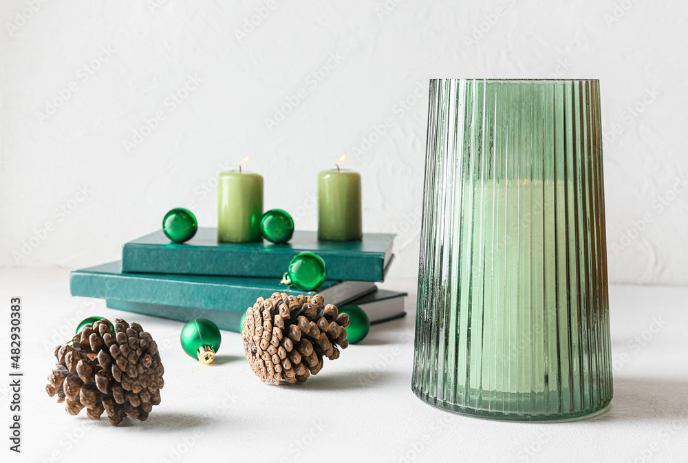 Holder with candle, pine cones and books on light background