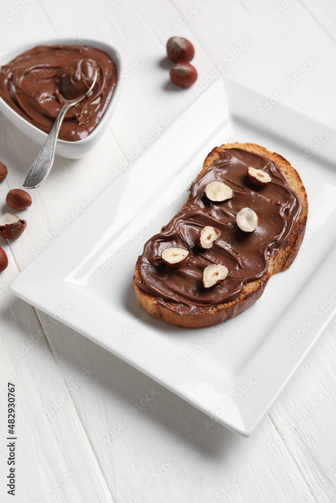 Plate of bread with chocolate paste and hazelnuts on white wooden background