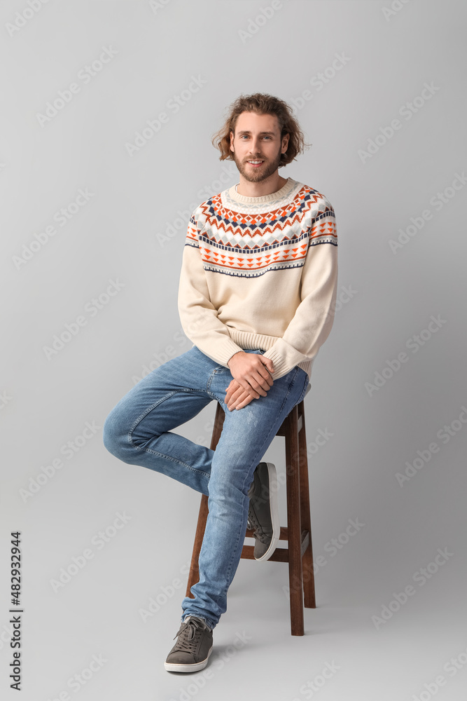 Handsome young man in knitted sweater sitting on chair against grey background