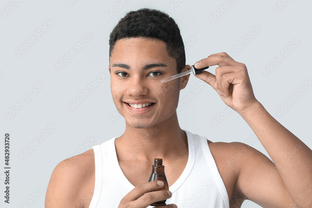 Young African-American guy with serum for skin care on grey background