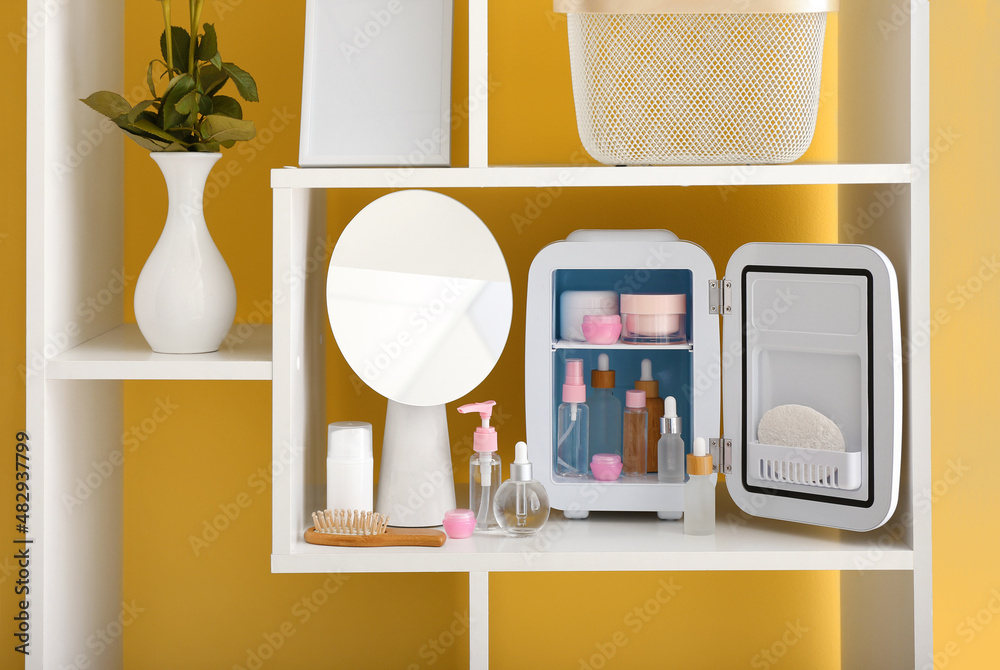 Shelf unit with small refrigerator and cosmetic products near color wall in room