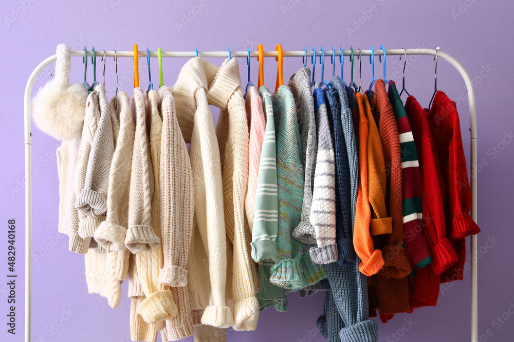 Rack with children sweaters and earmuffs near lilac wall