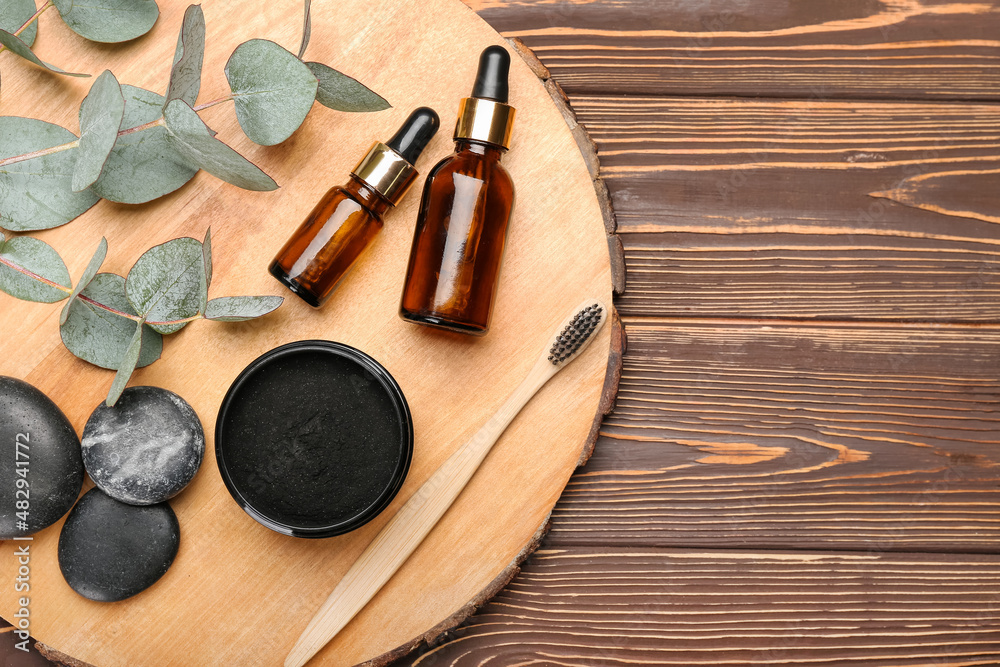 Jar with activated charcoal tooth powder, brush, cosmetic bottles and stones on wooden background