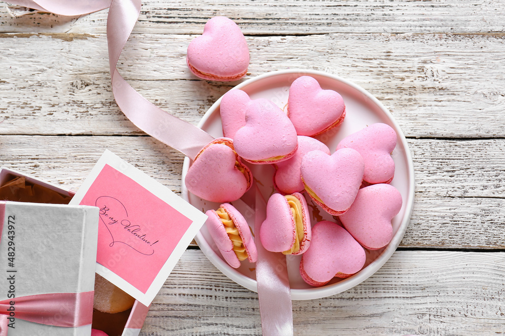 Plate with tasty heart-shaped macaroons and greeting card on white wooden background