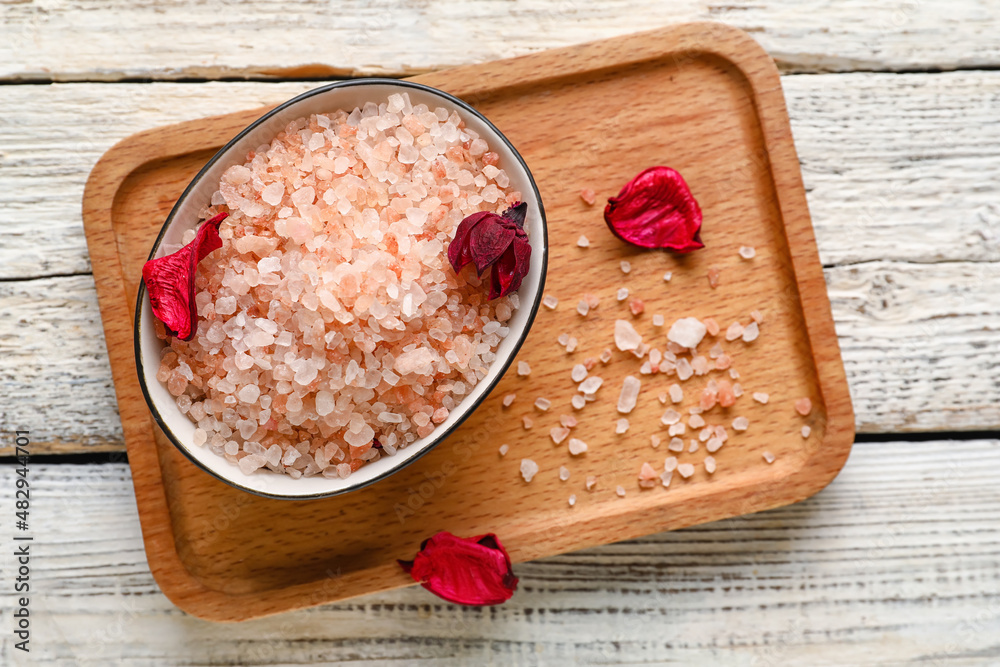 Board with bowl of sea salt and flower petals on light wooden background