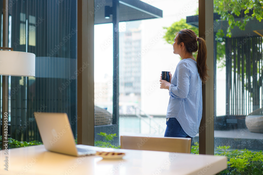 Asian woman drinking coffee in mug and smiling happily while taking a break from work at her compute