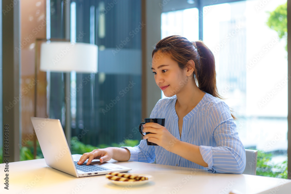 Asian woman drinking coffee in mug and smiling happily while taking a break from work at her compute