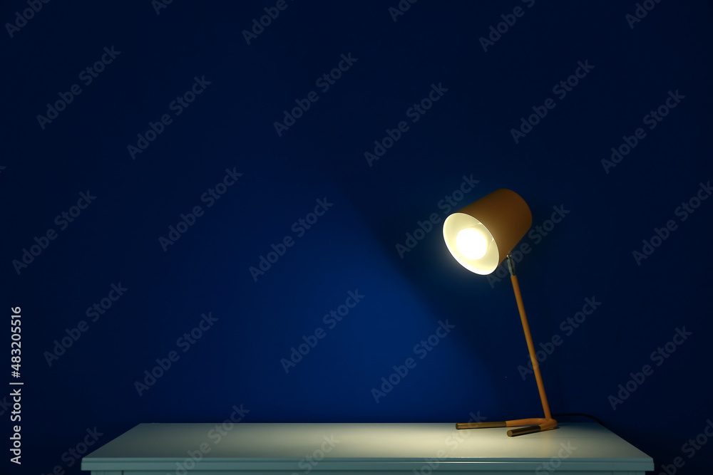 Glowing lamp on table near blue wall in dark room