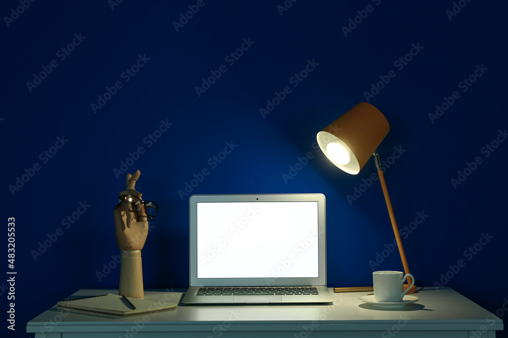 Modern workplace with open laptop and glowing lamp near blue wall in dark room