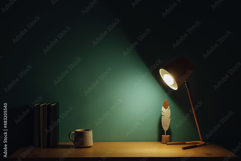 Glowing lamp, cup and books on table near green wall in dark room