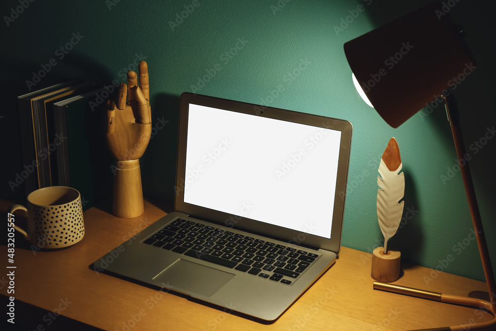 Modern workplace with open laptop and glowing lamp near green wall in dark room