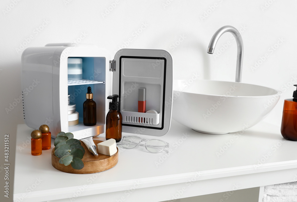 Open refrigerator with cosmetic products on table near light wall