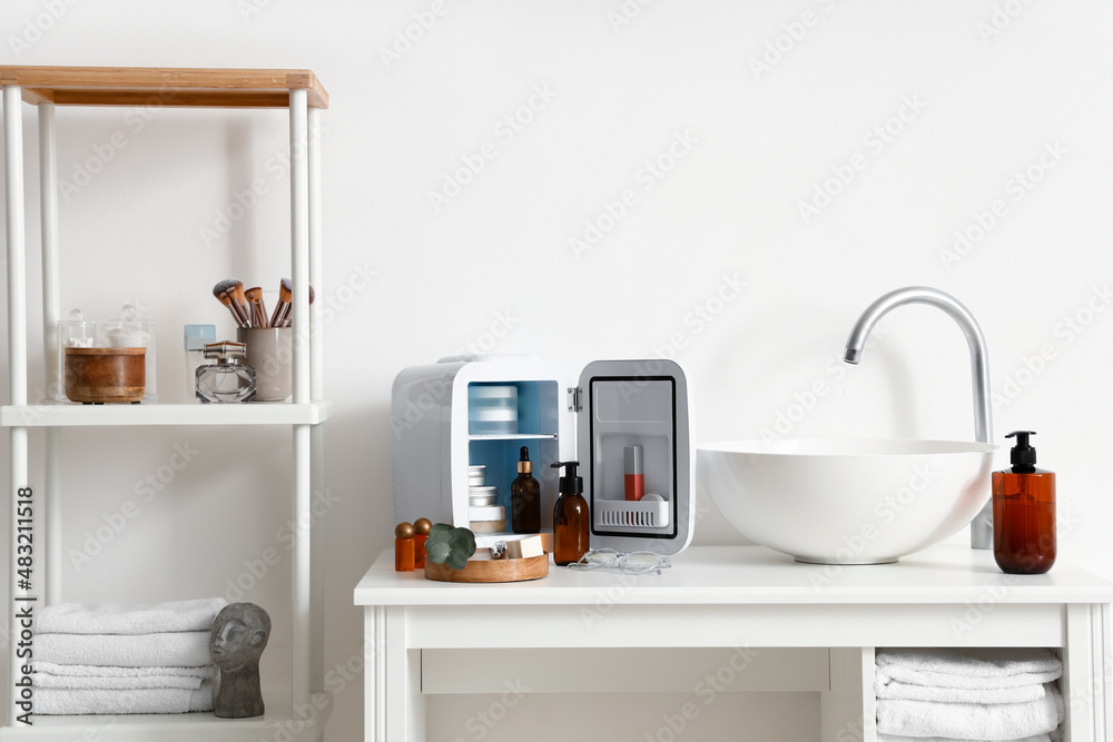 Sink, open refrigerator and cosmetic products on table near light wall in bathroom