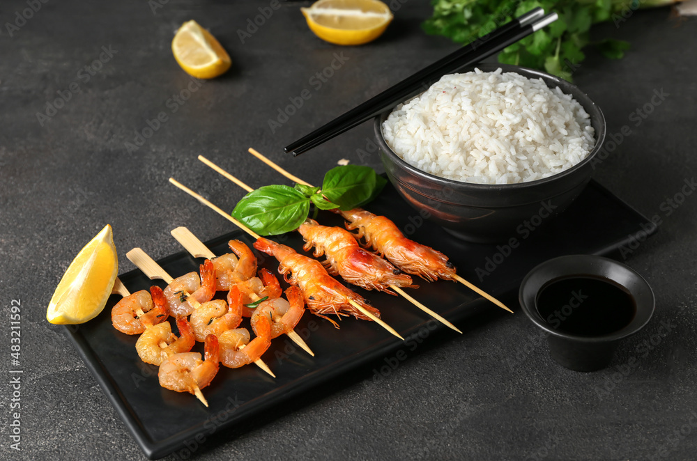 Board with tasty grilled shrimp skewers, sauce and rice on black background