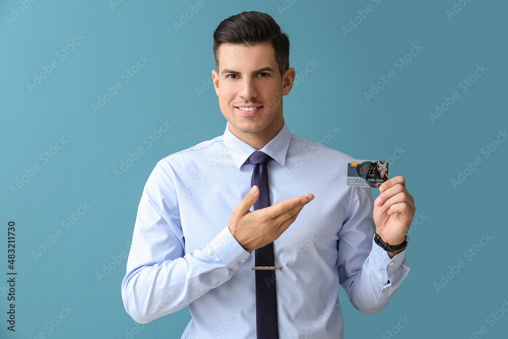 Handsome man in formal clothes pointing at gift card on color background