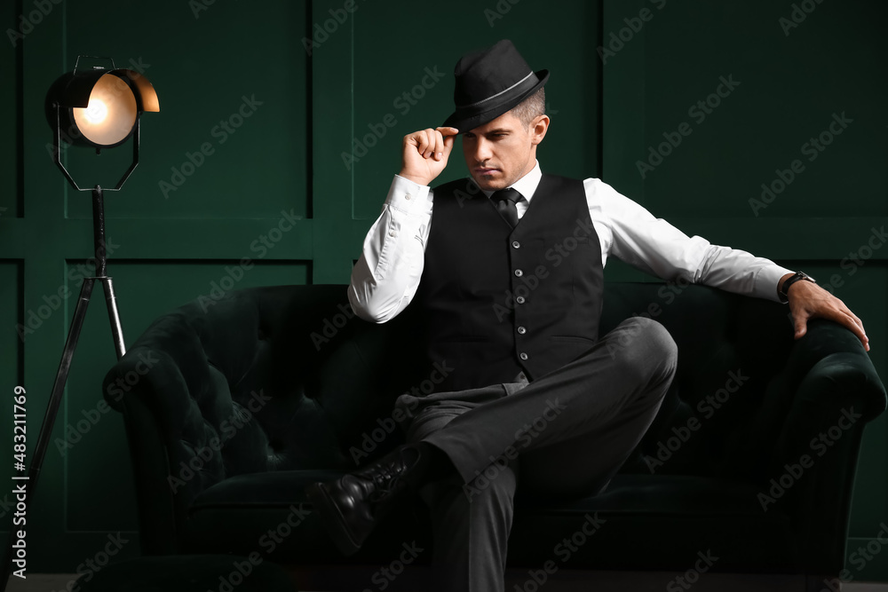 Gentleman in formal clothes and stylish hat sitting on sofa against color wall