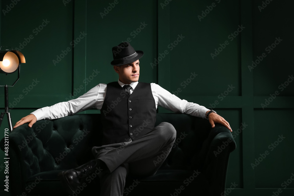 Gentleman in formal clothes and stylish hat sitting on sofa and looking aside against color wall