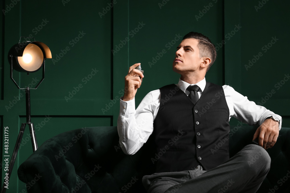 Gentleman in formal clothes sitting on sofa and spraying perfume from bottle near color wall