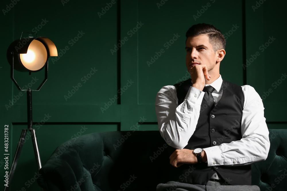 Pensive fashionable gentleman sitting in sofa near dark wall