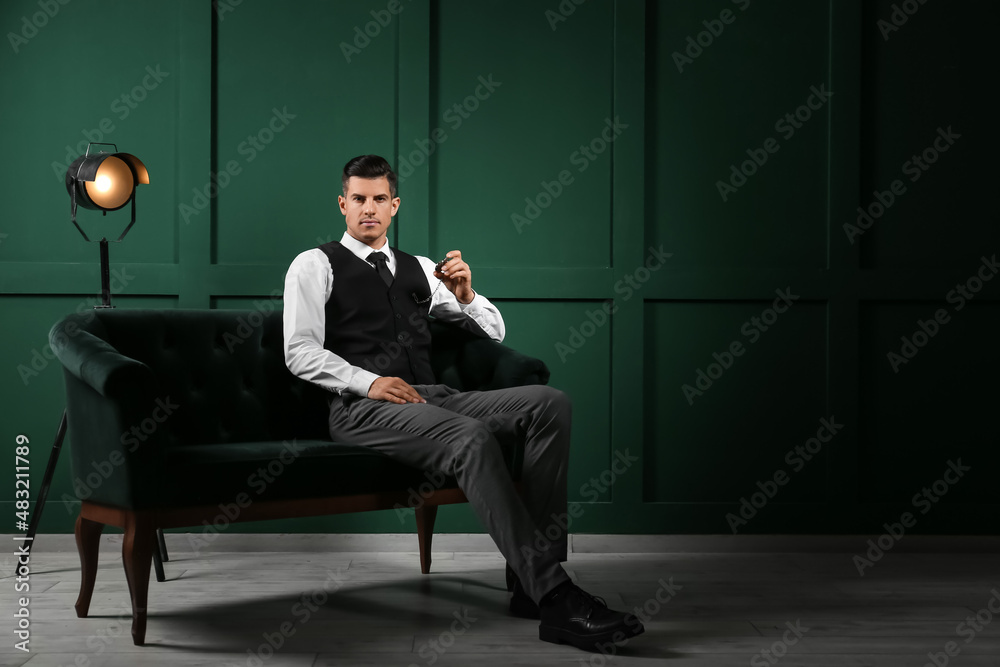 Fashionable gentleman sitting on sofa and holding watch in dark room