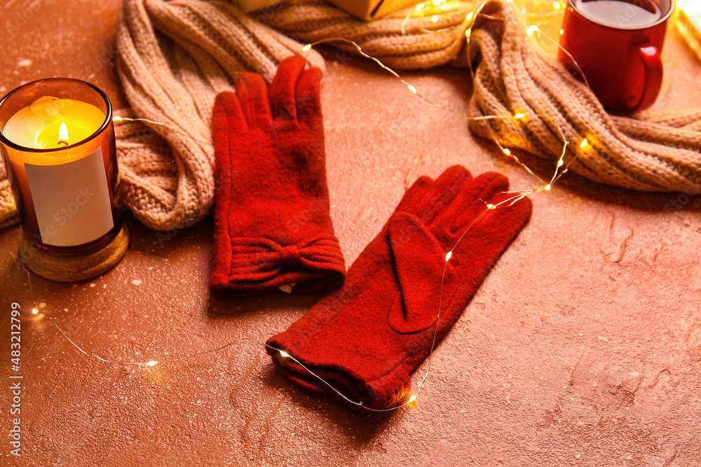 Warm gloves, scarf, Christmas lights and burning candle on red background