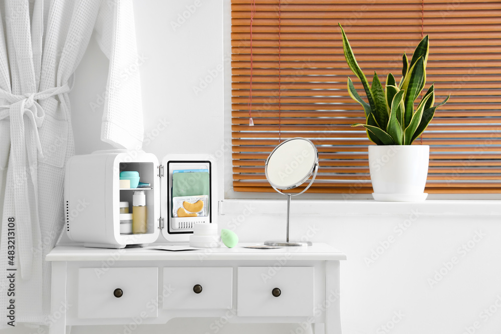 Small cosmetic refrigerator and mirror on dressing table near light wall