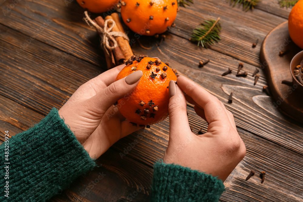 Woman making handmade Christmas decoration made of tangerine with cloves on wooden background, close