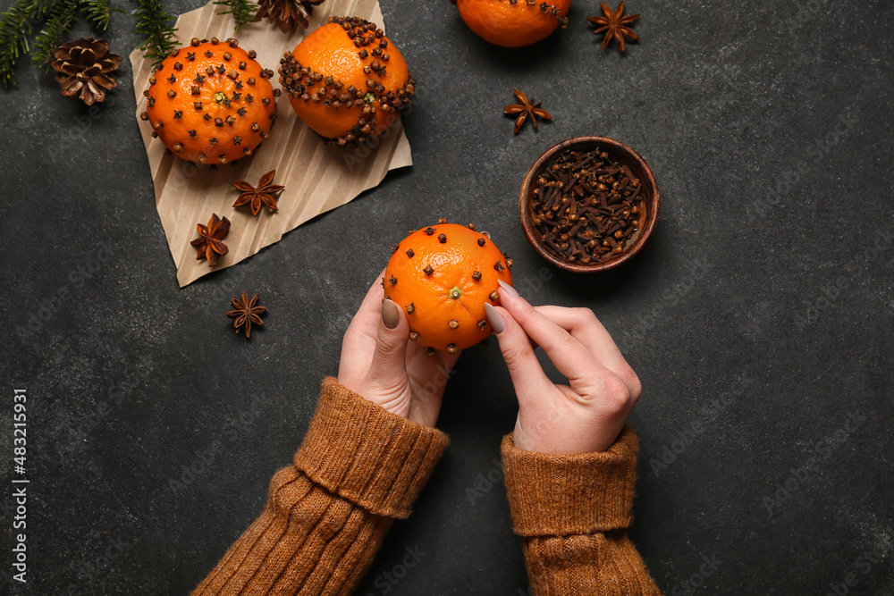 Woman making handmade Christmas decoration made of tangerine with cloves on dark background