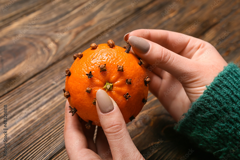 Woman making handmade Christmas decoration made of tangerine with cloves on wooden background, close