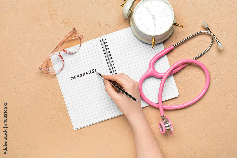 Woman writing word MENOPAUSE in notebook on beige background