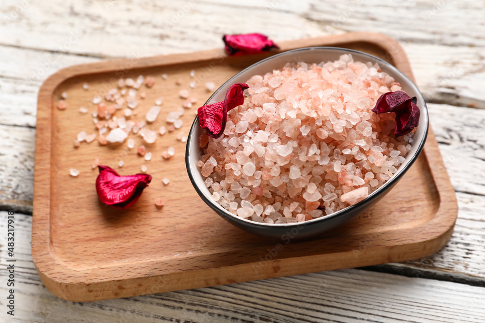 Board with bowl of sea salt and flower petals on light wooden background, closeup