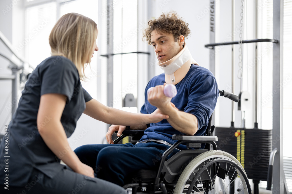 Rehabilitation specialist helps a guy to do exercises for recovery from injury, who is sitting in a 