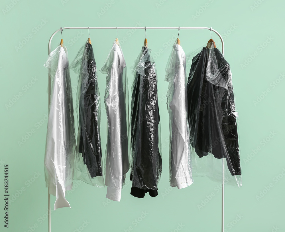 Rack with clean jackets and shirts in plastic bags on green background