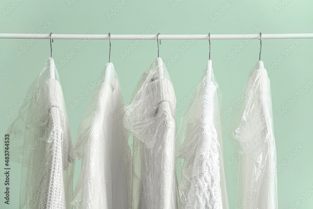 Rack with clean knitted sweaters in plastic bags on green background, closeup