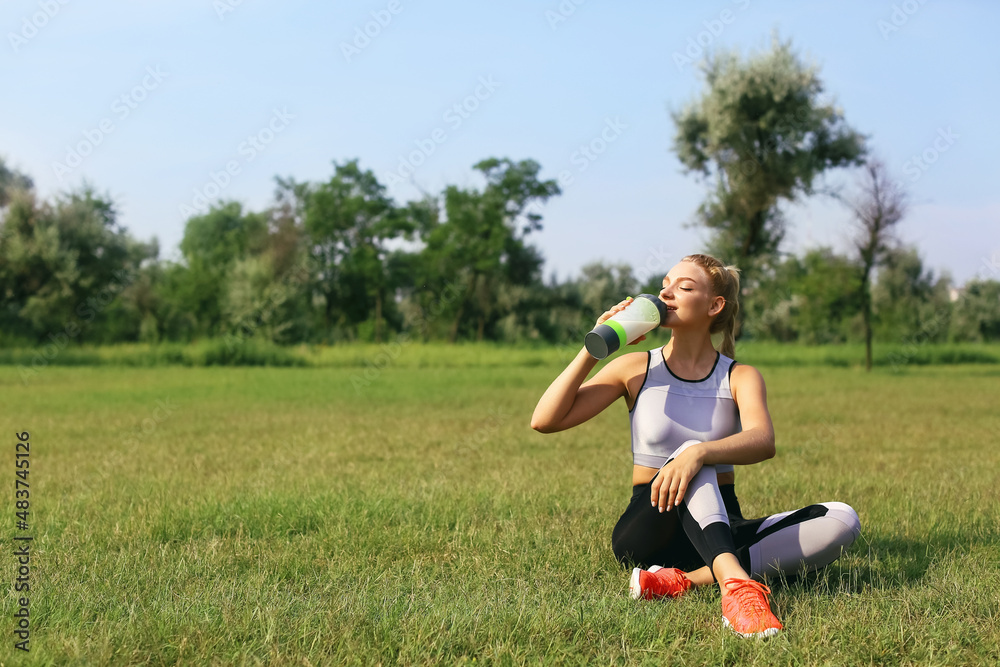 Young woman in sportswear sitting on grass and drinking protein shake