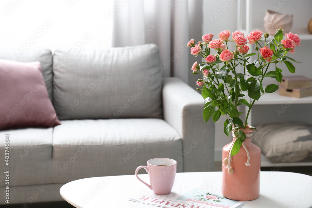 Vase with beautiful fresh roses and cup of coffee on table in room