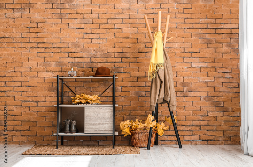 Hanger with jacket and basket with autumn leaves near brick wall in hallway