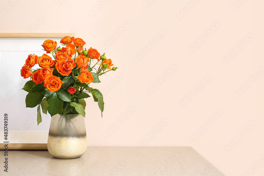 Vase with beautiful orange roses and blank photo frame on table against light wall