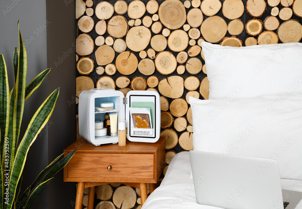 Small cosmetic refrigerator on bedside table near wooden wall