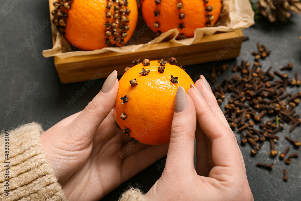 Woman making handmade Christmas decoration made of tangerine with cloves on dark background, closeup