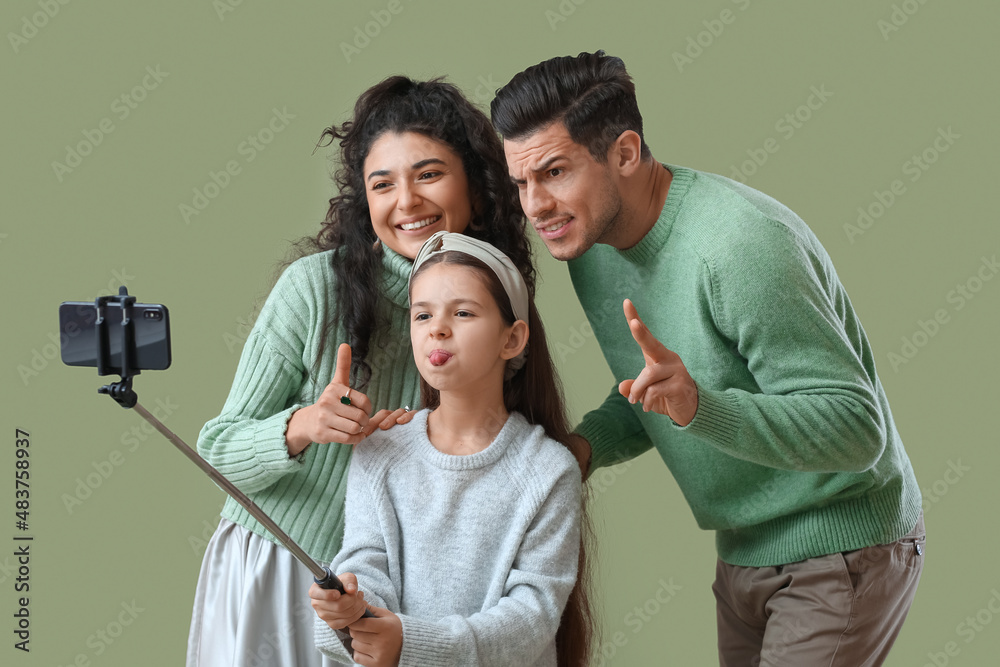 Happy parents with little daughter in warm sweaters taking selfie on green background