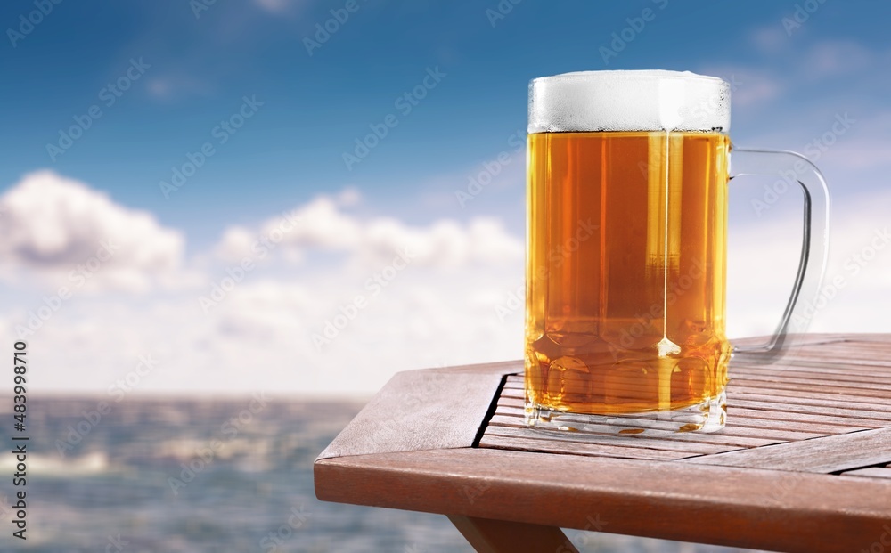 A glass of beer with condensation on the wooden table. Blurred sparkling sea at the background. Summ