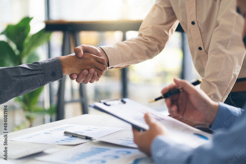 Entrepreneurs collaboration deal shaking hands in a modern office and financial paper graph on desk.