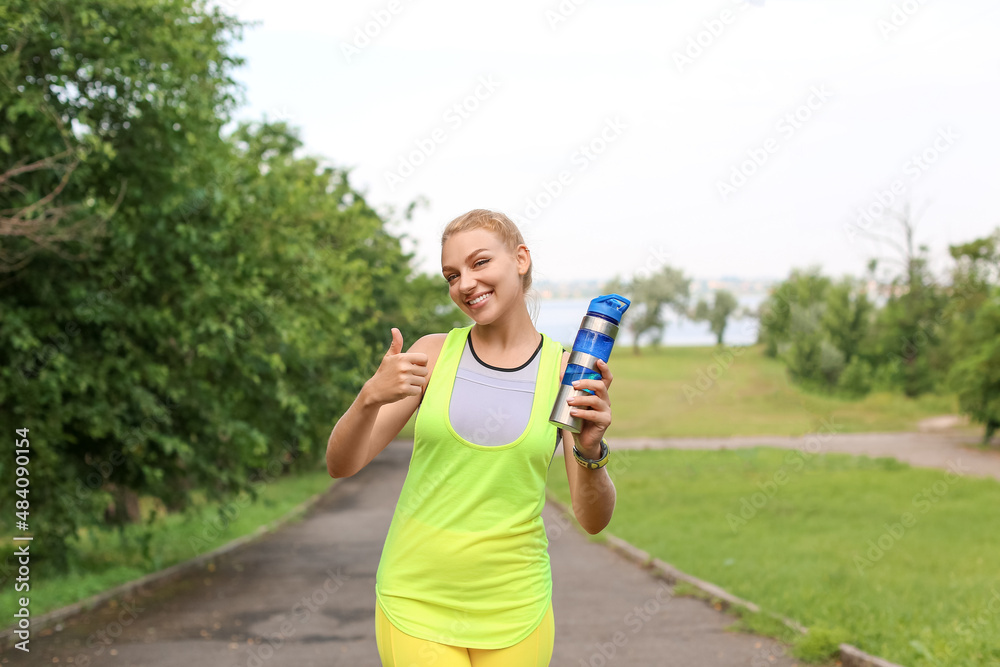 Young woman in sportswear with bottle of protein shake showing thumb up outdoors