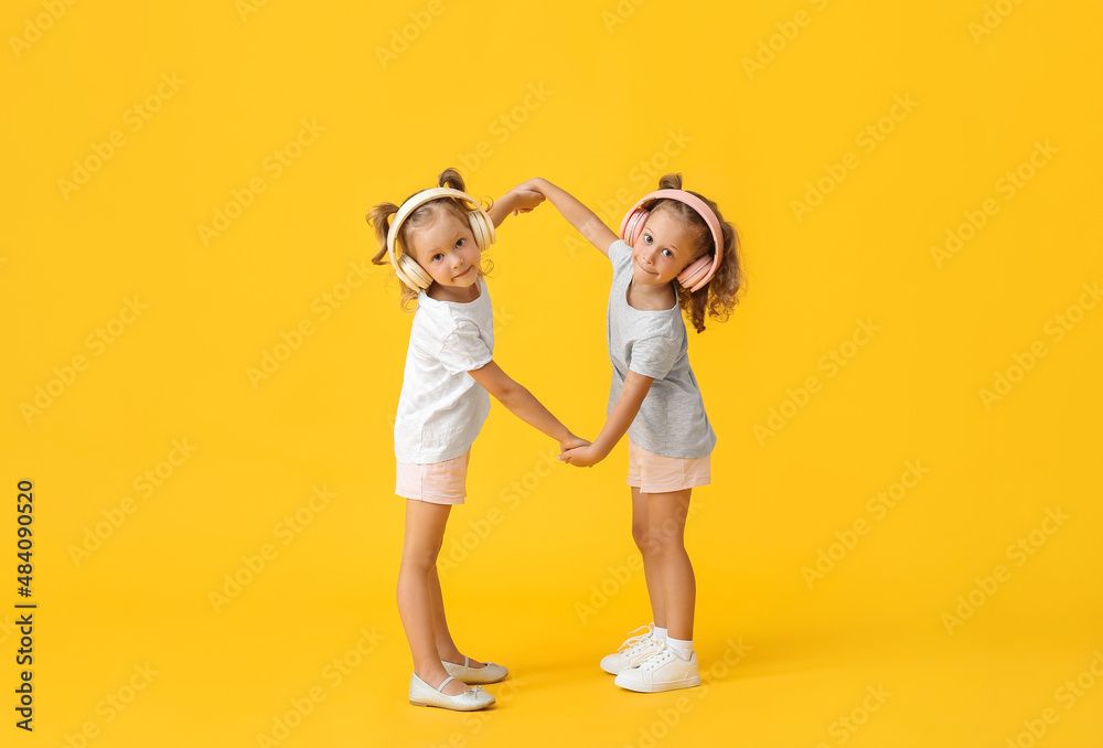 Cute little girls in headphones holding hands on yellow background