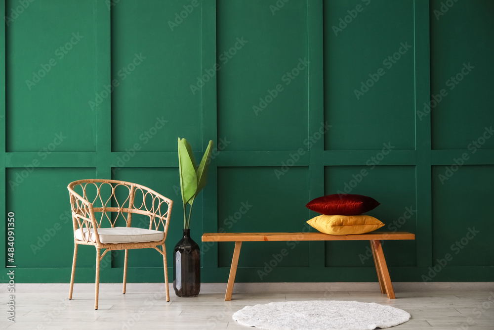 Wicker chair, vase with palm leaves and bench with pillows near color wall in room interior