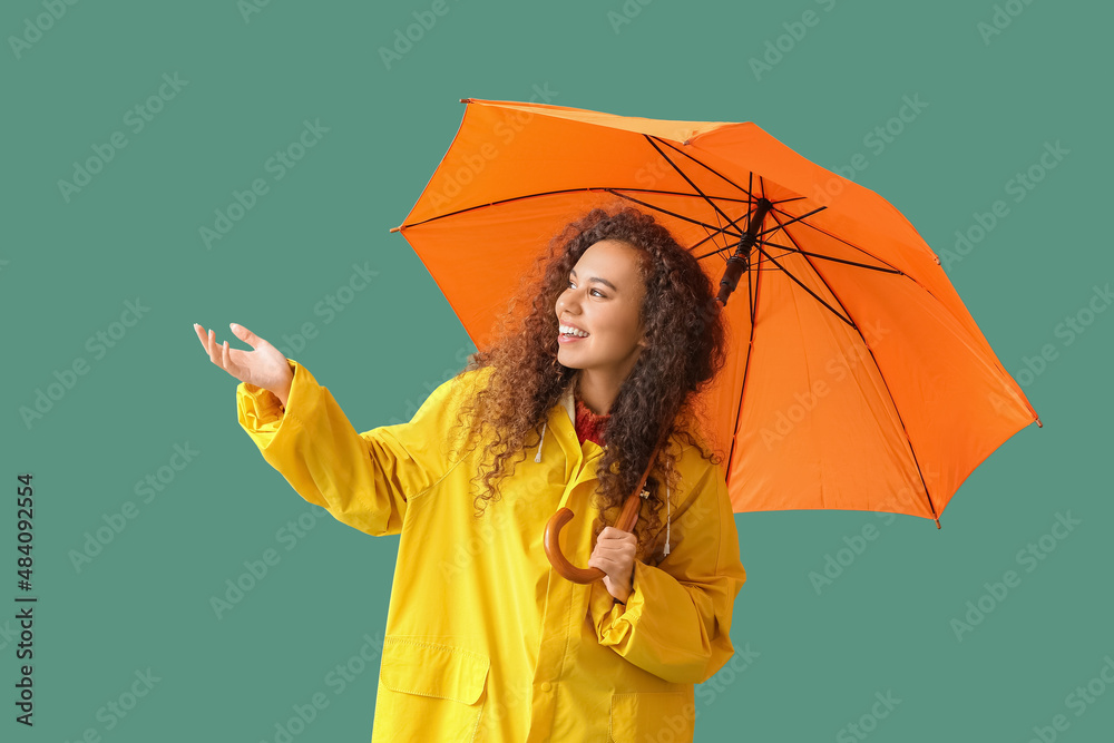 Young African-American woman in yellow raincoat with umbrella on green background