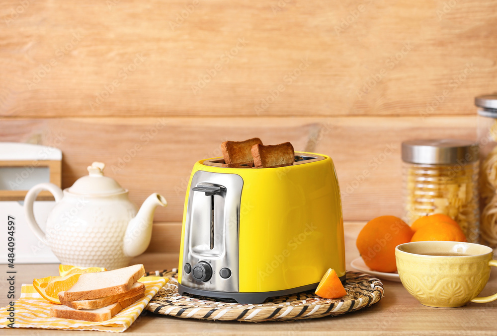 Yellow toaster with bread slices, oranges and cup of tea on table near wooden wall