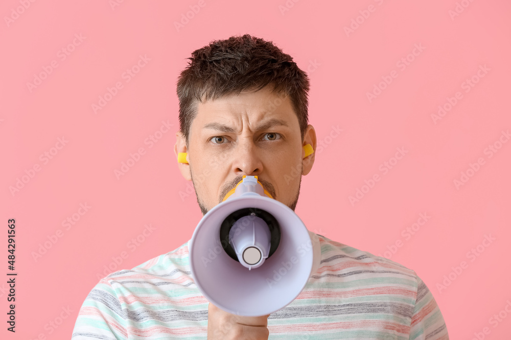 Angry young man with ear plugs shouting into megaphone on pink background