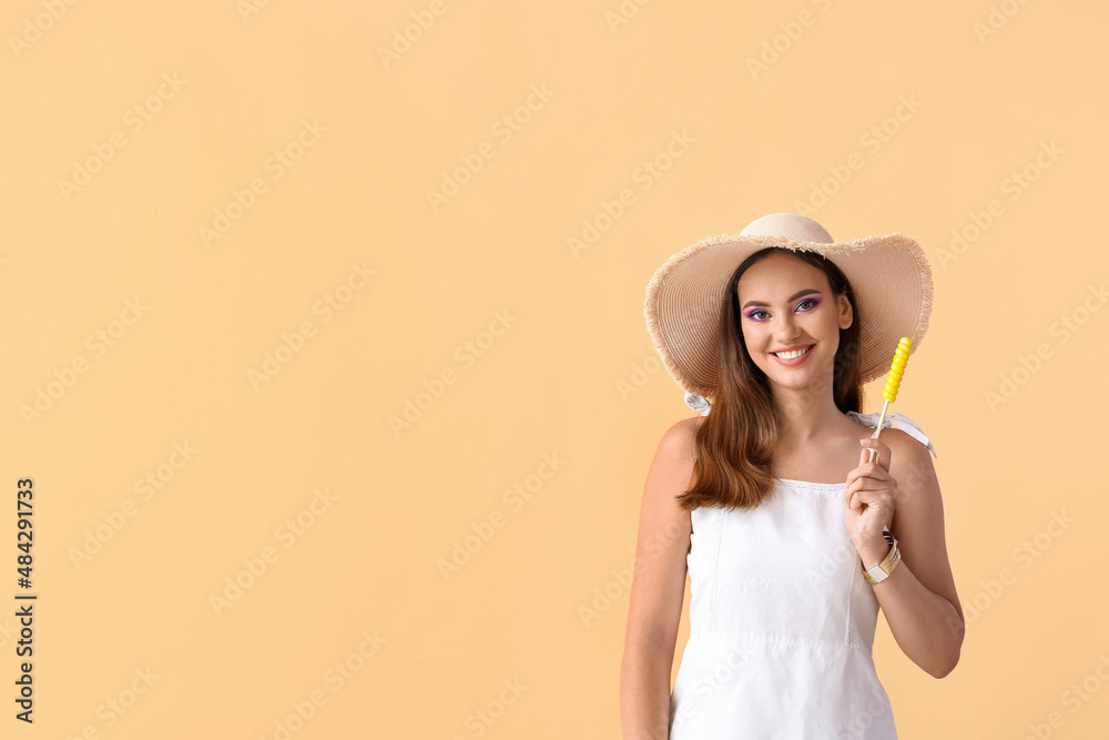 Beautiful young woman in hat with sweet lollipop on beige background